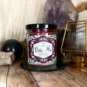 Op de afbeelding: Een rode glazen potkaars met een zwarte deksel en een etiket met de tekst "Le'bleu Apothecary Bite Me Offering | Ritual Parasoy Candle Net Wt 8 Oz | 227 g".