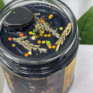 May include: Black candle with gold glitter and dried herbs in a glass jar. The label reads "L'eblou Apothecary" and "Ancestral Protection Oil of The Dead".