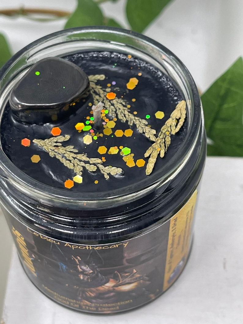 May include: Black candle with gold glitter and dried herbs in a glass jar. The label reads "L'eblou Apothecary" and "Ancestral Protection Oil of The Dead".
