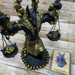 May include: A black and gold Anubis statue with a scale in each hand. The statue is surrounded by skulls and is sitting on a wooden surface. The statue is holding a heart in one scale and a feather in the other. The statue is a representation of the Egyptian god of the dead and the underworld. The image also includes a small package of soy wax melts with the text "Le'blet Apothecary" and "Anubis God of the dead & the underworld Soy Wax Melts Net wt. 3 Oz".
