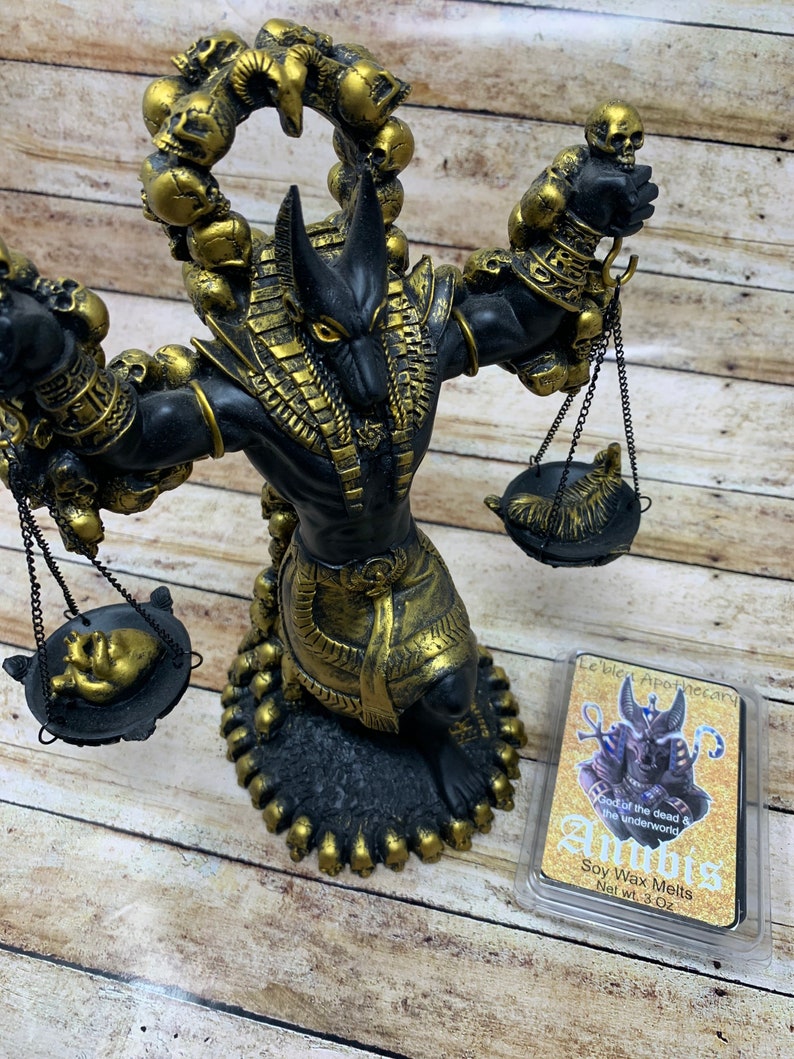 May include: A black and gold Anubis statue with a scale in each hand. The statue is surrounded by skulls and is sitting on a wooden surface. The statue is holding a heart in one scale and a feather in the other. The statue is a representation of the Egyptian god of the dead and the underworld. The image also includes a small package of soy wax melts with the text "Le'blet Apothecary" and "Anubis God of the dead & the underworld Soy Wax Melts Net wt. 3 Oz".