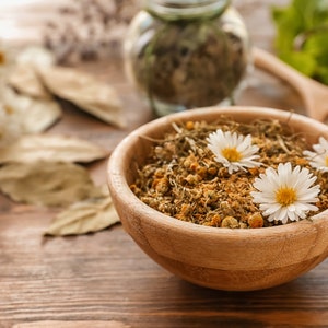 May include: A wooden bowl filled with dried chamomile flowers and a few fresh chamomile flowers on top. The bowl is on a wooden table with other dried herbs and flowers around it.