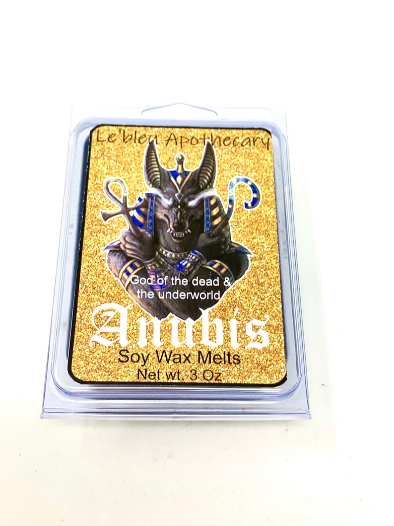May include: A gold and black wax melt container with an image of Anubis, the Egyptian god of the dead. The container is labeled "Le'blen Apothecary" and "Anubis Soy Wax Melts Net wt. 3 Oz".