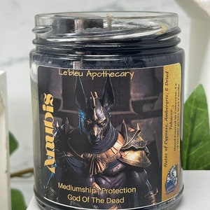 May include: A black glass candle jar with a gold label featuring an image of Anubis, the Egyptian god of the dead. The label also includes the text "Mediumship Protection God of The Dead" and "Le'bleu Apothecary". The candle has notes of cypress, ambergris, and dried tobacco.
