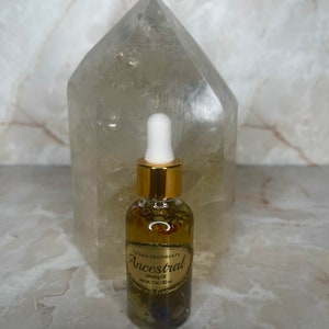 Op de afbeelding: Een klein glazen flesje met een gouden dop en een druppelaar. Het flesje is gevuld met een heldere vloeistof en bevat gedroogde kruiden. Het etiket vermeldt "Le Bleu Apothecary Ancestral Offering Oil Net Wt. 1 oz (30 ml)"