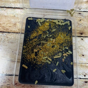 May include: A black wax melt with gold glitter and green botanicals in a clear plastic container.