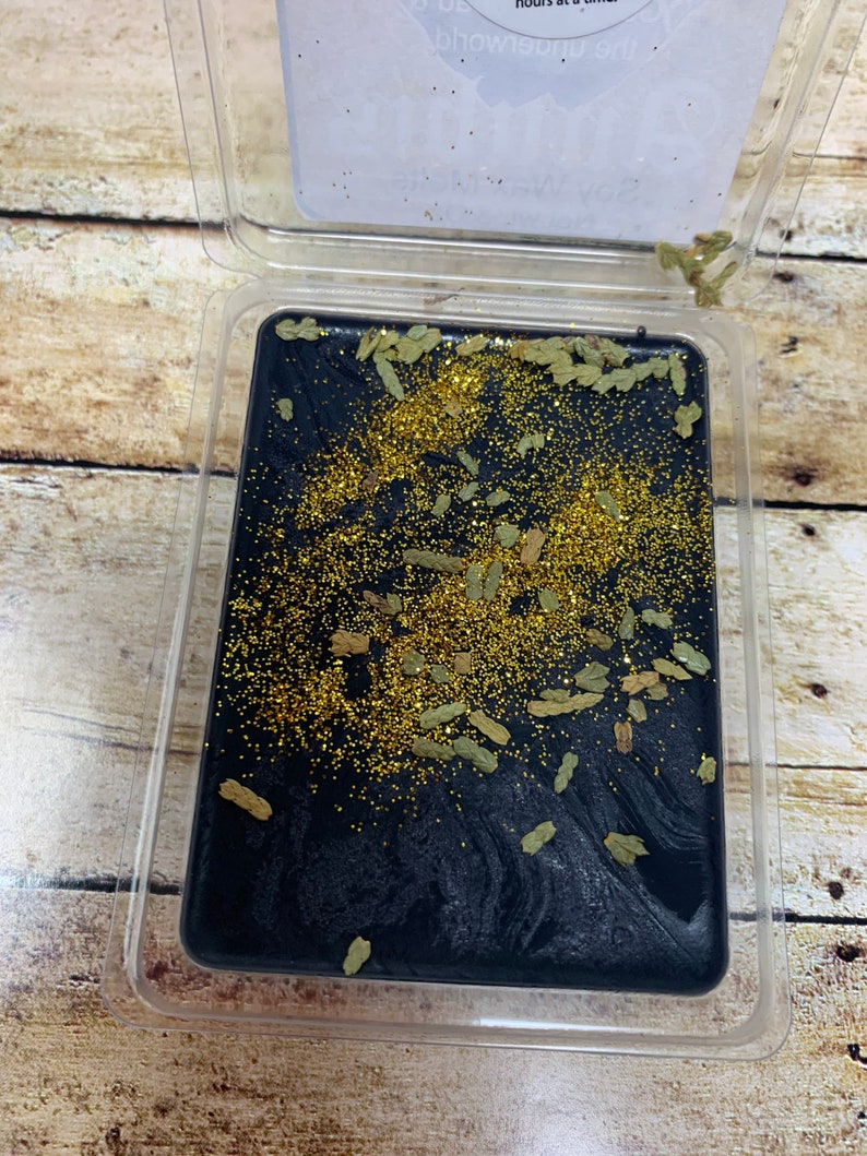 May include: A black wax melt with gold glitter and green botanicals in a clear plastic container.