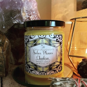 May include: A yellow candle in a glass jar with a black lid. The label reads "Le'bleu Apothecary Solar Plexus Chakra Offering Ritual Parasoy Candle Net Wt 8 oz | 227 g". The candle is surrounded by crystals and a gold candle holder.