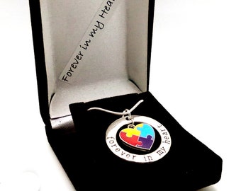 May include: A silver necklace with a heart-shaped pendant featuring a colorful puzzle piece design. The pendant is inscribed with the words "forever in my heart". The necklace is displayed in a black velvet box.