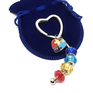 May include: A silver heart-shaped keychain with a colorful puzzle piece charm and a string of colorful beads. The keychain is in a blue velvet pouch.