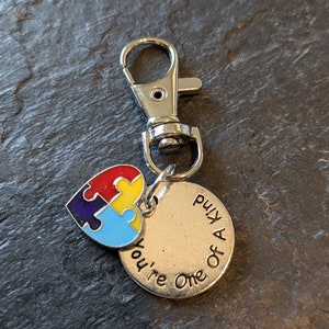 May include: Silver keychain with a blue, yellow, red, and purple puzzle piece heart charm. The charm is attached to a round silver tag that reads "You're One of a Kind".