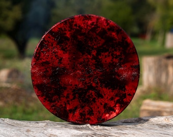 Puede incluir: Un objeto redondo con un patrón rojo y negro, posiblemente un parche de tambor, descansa sobre un tronco desgastado. El diseño presenta una combinación de colores rojo oscuro y negro. El fondo está borroso, mostrando follaje verde.