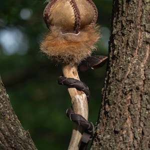 May include: A decorative staff featuring a curved antler handle wrapped in dark brown leather cord. The top consists of a rounded, tan fabric sphere accented with brown stitching and surrounded by a thick layer of textured, light brown fur.
