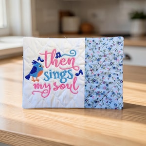 May include: A quilted fabric item with the embroidered text "Then sings my soul" and a blue bird. The left side is white with a diamond pattern, and the right side has a blue floral pattern.