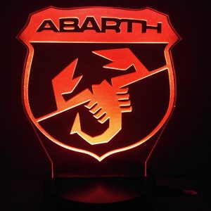 May include: A red illuminated sign with the word "ABARTH" in black lettering and a scorpion logo. The sign is shaped like a shield.