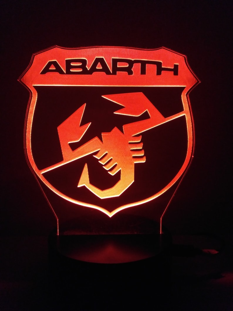 May include: A red illuminated sign with the word "ABARTH" in black lettering and a scorpion logo. The sign is shaped like a shield.