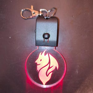 Puede incluir: Un llavero negro con un cierre plateado y un colgante redondo rojo brillante. El colgante presenta un diseño estilizado de cabeza de león naranja claro. El llavero tiene un botón rectangular negro y dos líneas rojas debajo.