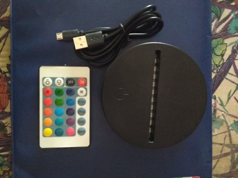 May include: A black round base with a white light strip and a remote control with buttons for different colors, white light, flash, smooth, fade, and mode. The remote control also has buttons for on and off.