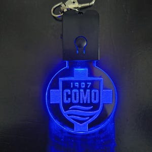Può includere: Un portachiavi nero con un moschettone argentato e un emblema blu luminoso. L'emblema presenta il testo "1907 COMO" all'interno di un design a scudo. Il portachiavi è illuminato da una luce blu brillante.