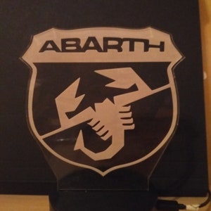 May include: A black and white night light featuring the Abarth logo. The logo is a shield with the word "ABARTH" at the top and a scorpion inside the shield.