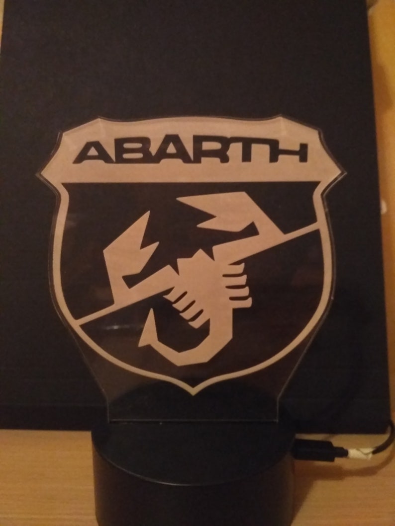 May include: A black and white night light featuring the Abarth logo. The logo is a shield with the word "ABARTH" at the top and a scorpion inside the shield.