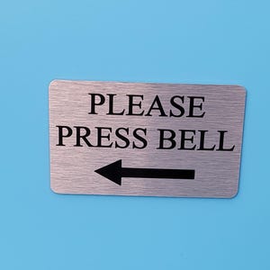 May include: A rectangular metal sign with the words "PLEASE PRESS BELL" in black, and a black arrow pointing left. The sign has a brushed metal finish and is set against a light blue background.