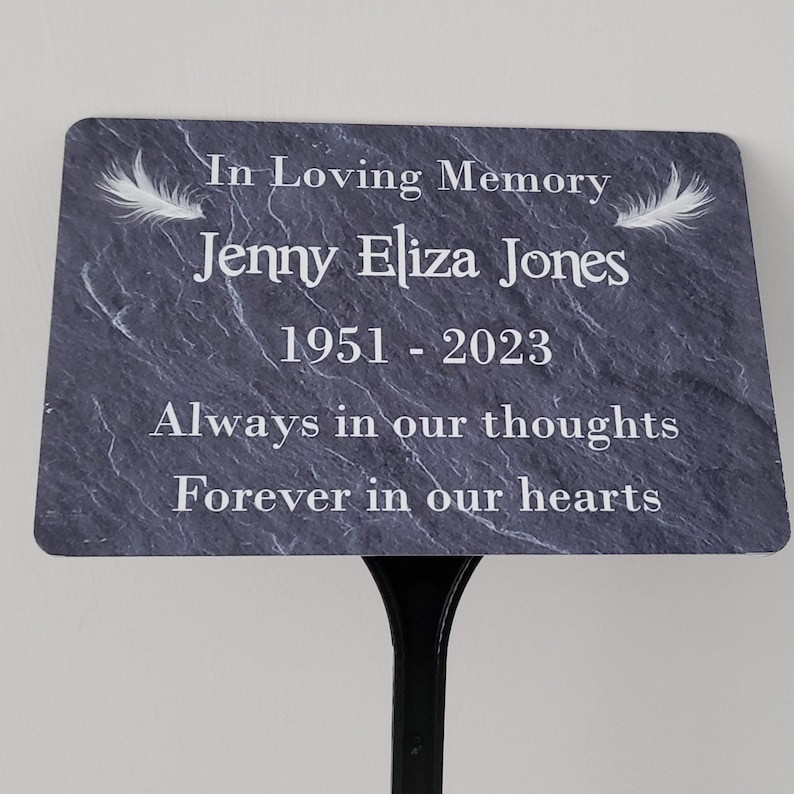 May include: A black slate memorial plaque with white text that reads "In Loving Memory Jenny Eliza Jones 1951-2023 Always in our thoughts Forever in our hearts". The plaque has two white feathers on either side of the text.