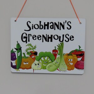 May include: A white metal sign with the text "Siobhann's Greenhouse" in black lettering. The sign features a cartoon illustration of various vegetables with faces, including a cucumber, a pumpkin, a carrot, a broccoli, a pepper, and an eggplant.