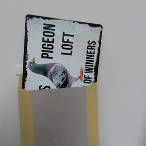 May include: A white metal sign with black text that reads "Pigeon Loft Home of Winners". A white pigeon with black and gray markings is pictured on the sign.