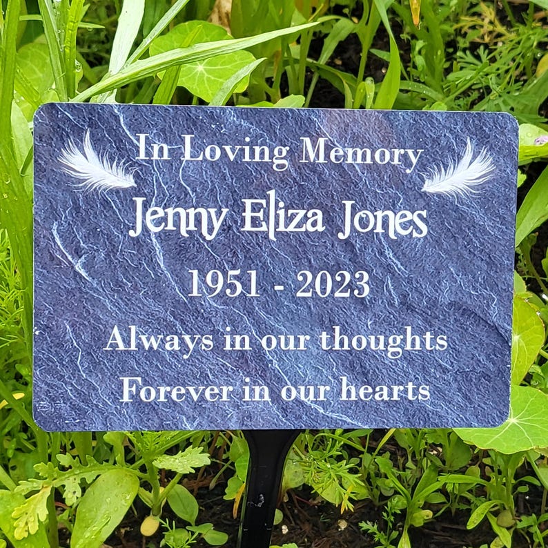 May include: A blue slate memorial plaque with white feathers and the text "In Loving Memory Jenny Eliza Jones 1951-2023 Always in our thoughts Forever in our hearts".