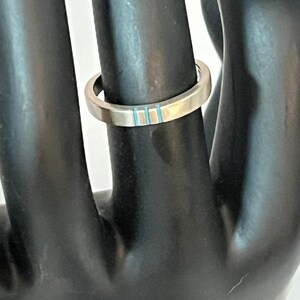 May include: A silver-toned metal ring featuring a flat band design with three vertical light blue inlay stripes positioned on the front surface. The ring has a smooth, polished finish and a consistent width throughout the entire circular band.