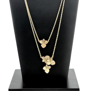Peut inclure: Un collier à double chaîne doré orné de breloques florales délicates. La chaîne la plus courte présente une fleur à trois pétales, tandis que la chaîne plus longue comporte un groupe de fleurs à trois pétales disposées en cascade.