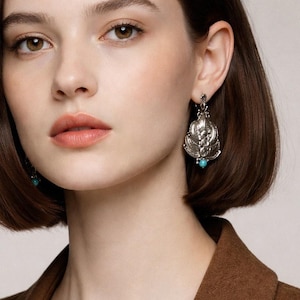 May include: A pair of silver-toned drop earrings featuring an ornate, filigree-style metal design with a central floral motif and a small, round, bright turquoise bead dangling from the bottom edge of each piece, secured with a hook closure.