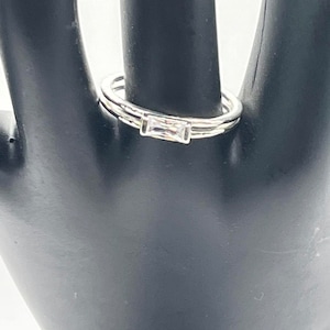 May include: A silver ring with a single clear rectangular stone set in the center. The ring is on a black mannequin hand.