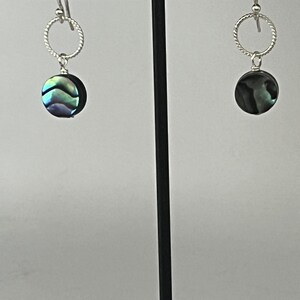 Può includere: Un paio di orecchini pendenti con dischi circolari in conchiglia di abalone dalle tonalità verdi e viola iridescenti. Ogni disco pende da un cerchio in filo ritorto argentato fissato a un semplice gancio argentato per l'orecchio.