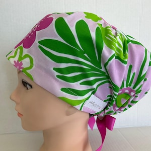 May include: A scrub cap featuring a vibrant floral pattern with large green leaves and bright pink flowers on a light lavender background. The cap includes a small white fabric tag with the text phillyfabrics and a pink ribbon tie at the back.