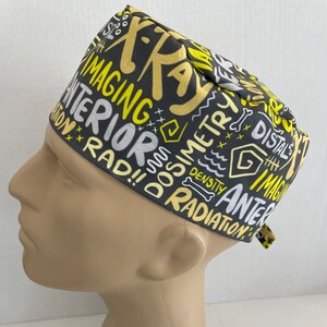 May include: A gray and yellow scrub hat with a variety of medical terms printed on it, including "X-ray", "Imaging", "Interior", "Radiation", "Dosimetry", "Density", "Distal", and "Anterior".