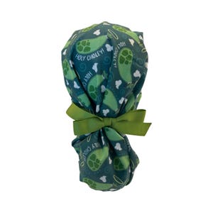 May include: A teal scrub cap with a pattern of green artichokes and white clouds. The words "Holy Choley!" are printed on the cap. A green ribbon is tied in a bow at the base of the cap.