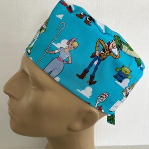 May include: A bright blue scrub cap featuring a repeating pattern of Toy Story characters including Woody, Bo Peep, Forky, and a green alien against a background of white clouds. The cap has a fitted crown and ties at the back for an adjustable fit.