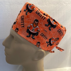 May include: Orange and black surgical scrub hat with a repeating pattern of Gritty, the Philadelphia Flyers mascot. The hat has a tie at the back and the text "Gritty" and "NHL" are printed on the hat.