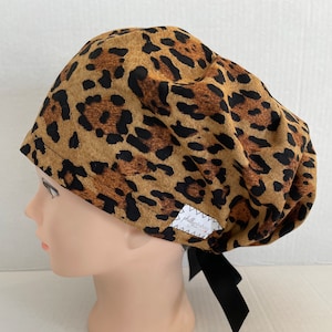 May include: A brown and black leopard print surgical scrub hat with a black tie. The hat has a white label with the text "philly scrubbers" on it.