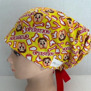 May include: Yellow surgical scrub hat with a red and white pattern featuring the game Operation. The hat has a red tie.