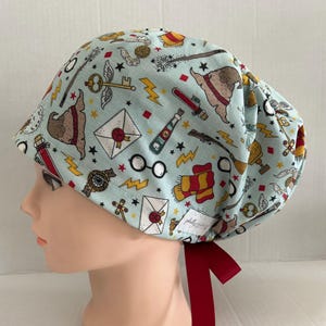 May include: A light blue patterned scrub cap featuring various magical themed illustrations. The cap includes images of a golden snitch, lightning bolts, and a sorting hat. A red ribbon is tied at the back of the cap.