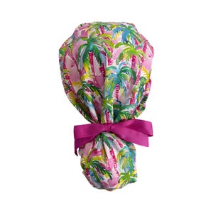May include: A pink scrub cap with a tropical palm tree pattern in shades of green, blue, and yellow. The cap is tied with a bright pink bow. This item is designed for medical professionals.