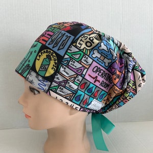 May include: A colorful surgical scrub cap with a variety of medical-themed graphics, including text that reads "LAPAROSCOPIC" and "OPERATING ROOM." The cap features a teal ribbon tie and is designed to be worn in an operating room setting.