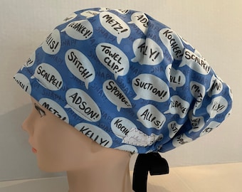 Könnte beinhalten: Blaue OP-Mütze mit einem Muster aus weißen Sprechblasen, die medizinische Begriffe wie "Skalpell!" und "Handtuchclip!" enthalten. Die Mütze hat eine schwarze Schleife.