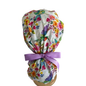 May include: A white scrub cap featuring a vibrant watercolor print of colorful hummingbirds and various wildflowers in shades of pink, orange, yellow, and purple. The cap is gathered at the back and secured with a solid light purple ribbon bow.