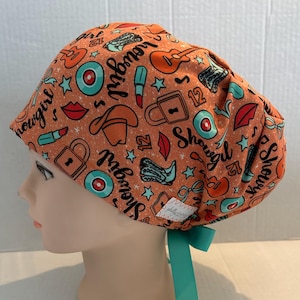 May include: An orange scrub cap with a teal ribbon tie, patterned with cowboy boots, guitars, lipsticks, and the word "Showgirl". The cap is designed for medical professionals and features a comfortable fit.