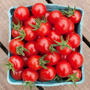 May include: A full container of ripe, red cherry tomatoes with green stems. The tomatoes are in a light blue paperboard container, set against a wooden surface. The tomatoes are round and shiny.