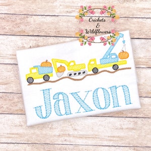 May include: A white onesie with an embroidered design of a yellow construction truck, excavator, and crane carrying pumpkins. The name "Jaxon" is embroidered in blue below the trucks.